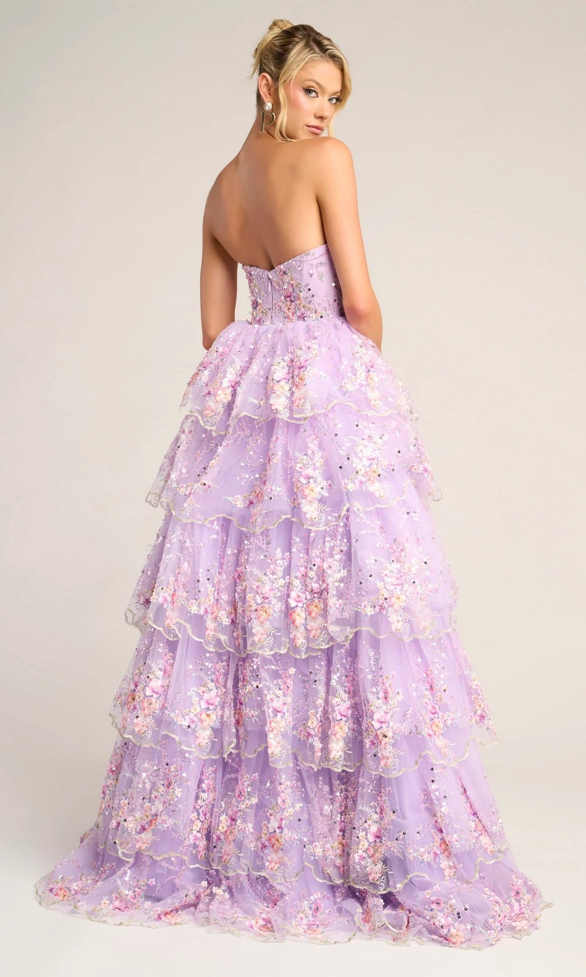 A woman poses with her back to the camera in the Portia and Scarlett PS26051 Strapless Floral Ball Gown, featuring tiered lavender layers and embroidered floral applique, against a neutral background.